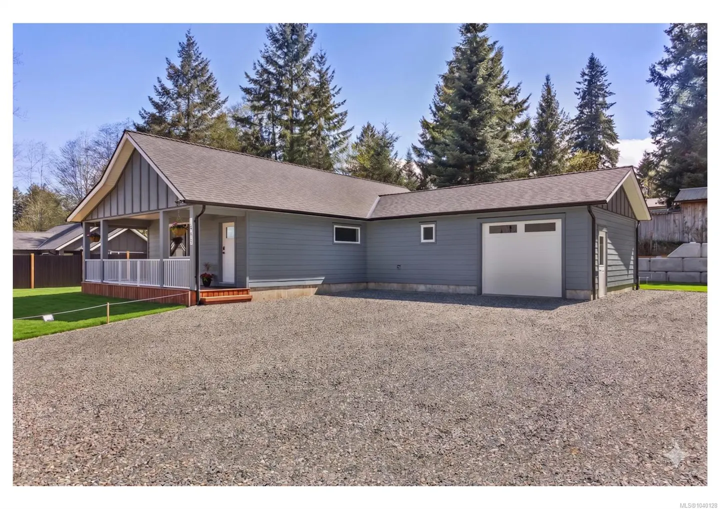 Single-story grey house with a covered porch, white garage door, and gravel driveway, surrounded by tall pine trees.