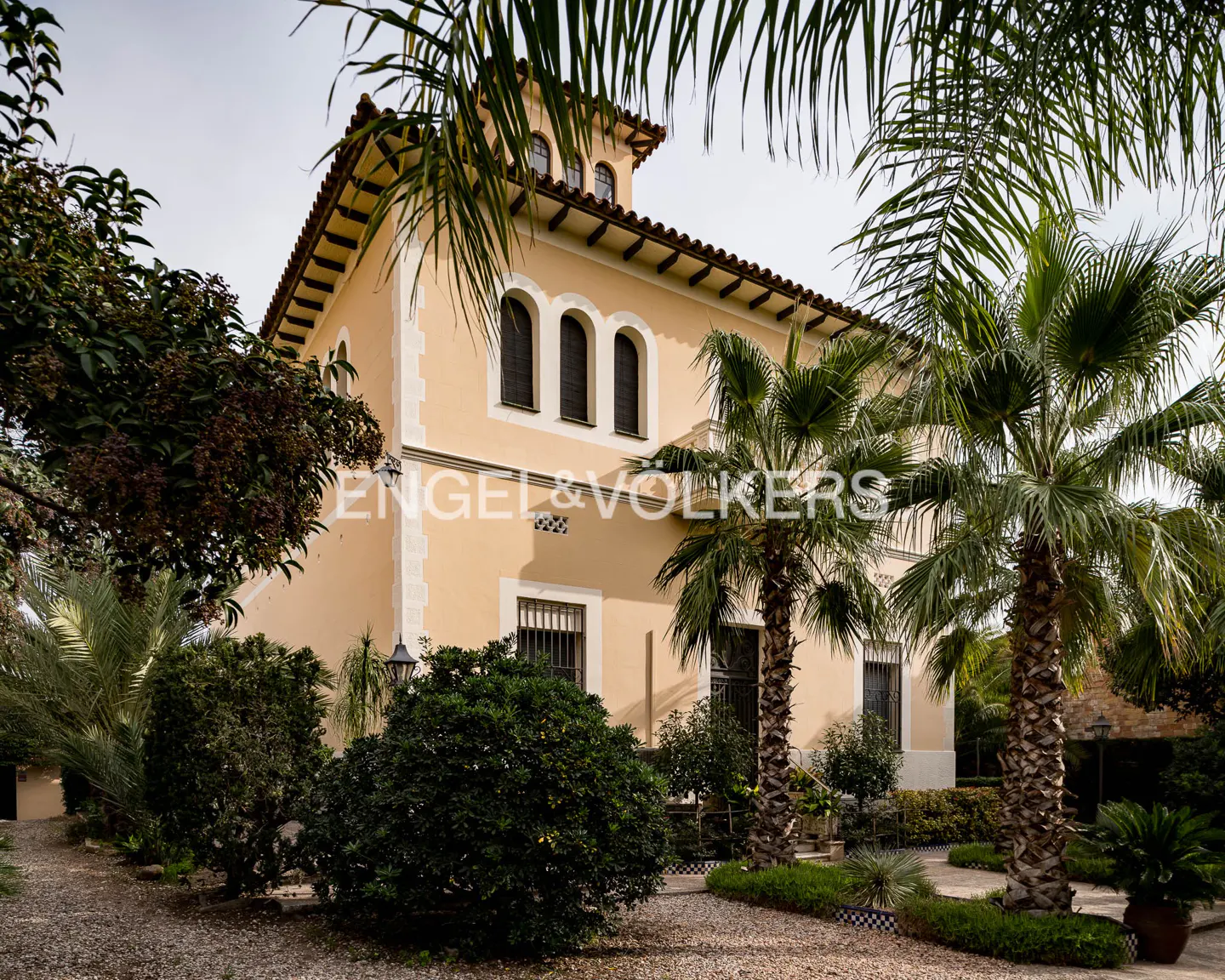 Exterior view of a two-story, light yellow house with palm trees and green bushes in the front yard.