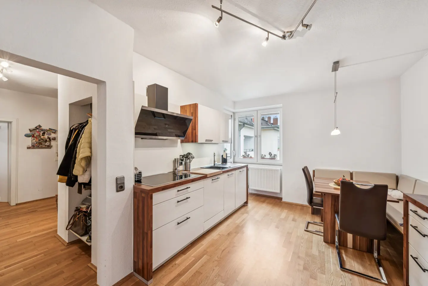 Bright kitchen with wood floors, white cabinets, and a dining area with a brown table and chairs. A coat closet is visible in the background.