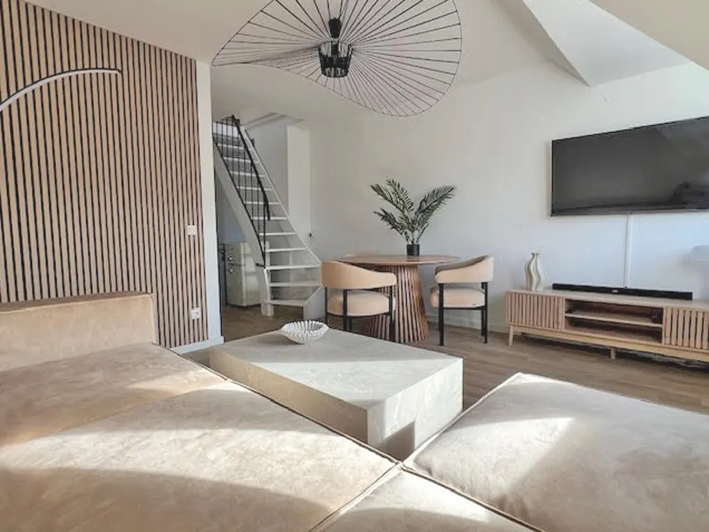 A bright living room with a beige couch, a stone coffee table, and a round wooden dining table with two chairs. A TV hangs on the white wall.