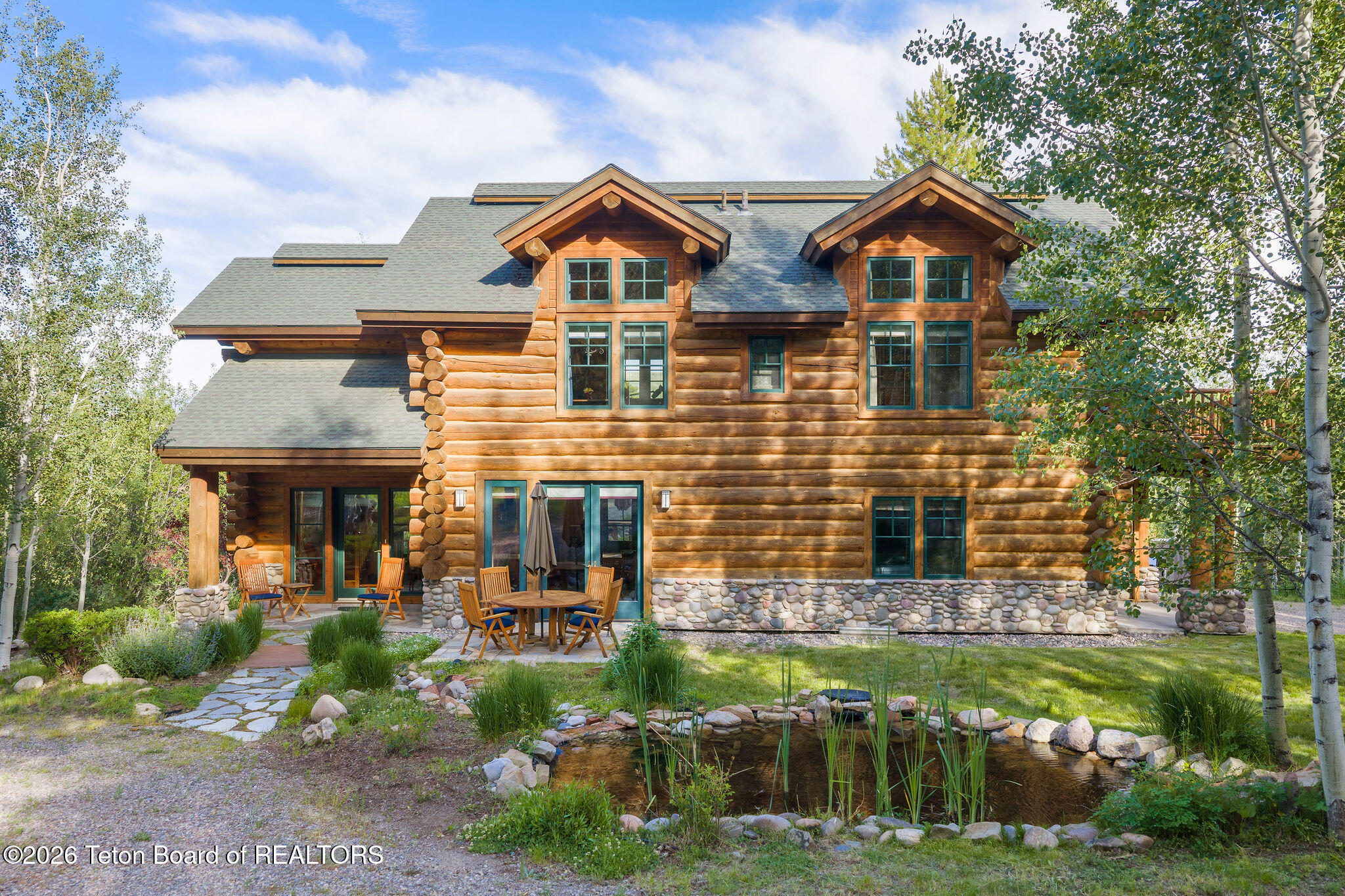 A large, two-story log cabin home with a stone foundation, patio furniture, and a small pond in a wooded yard.
