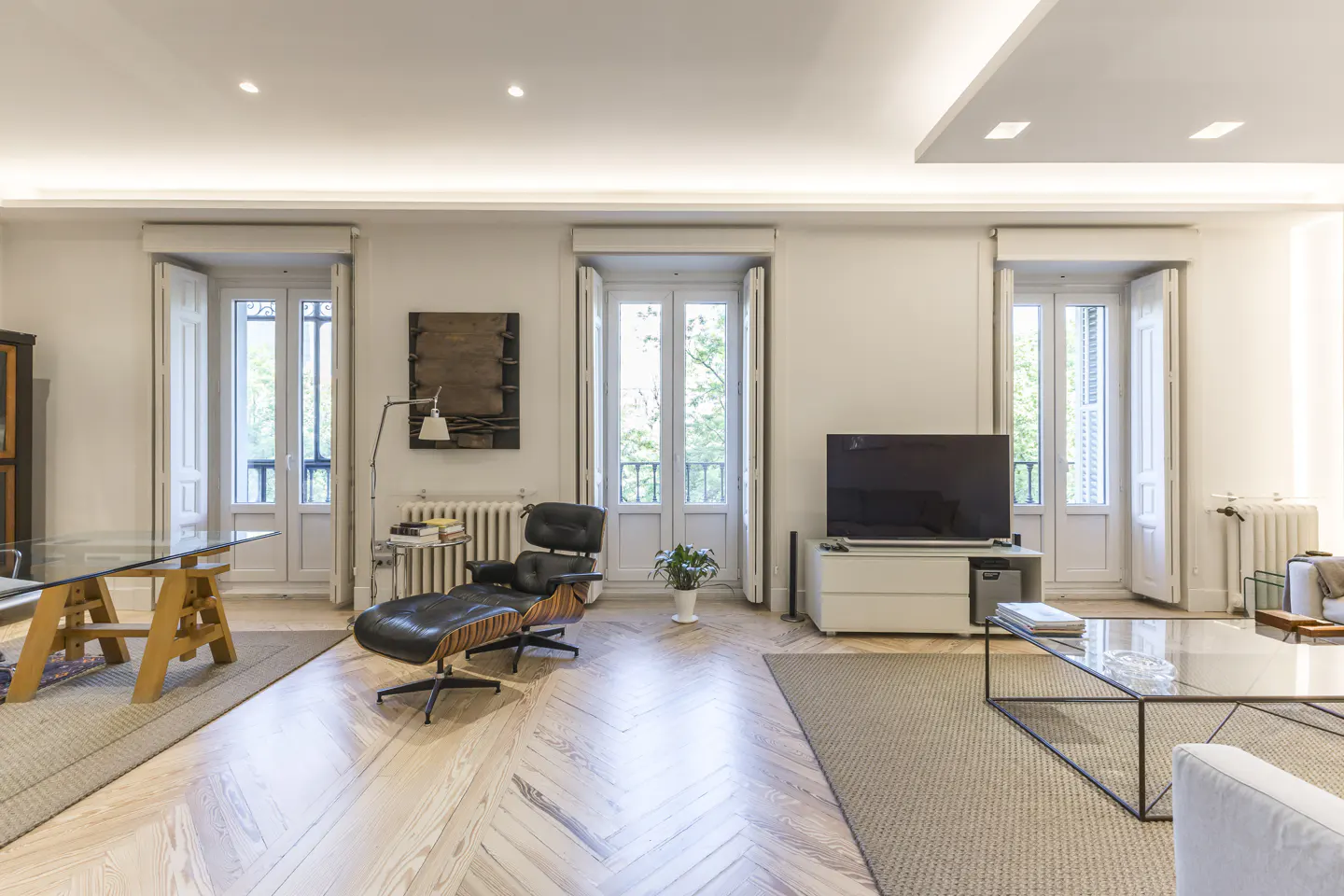 Bright living room with herringbone wood floors, white walls, and three sets of French doors. A black leather chair and TV are visible.