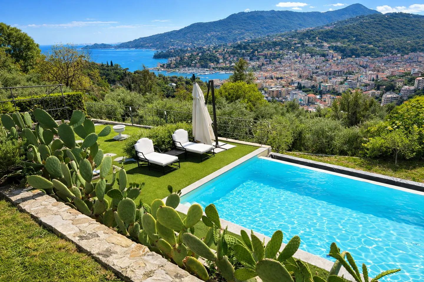 A bright blue pool with two white lounge chairs and a white umbrella overlooks a city and the ocean.
