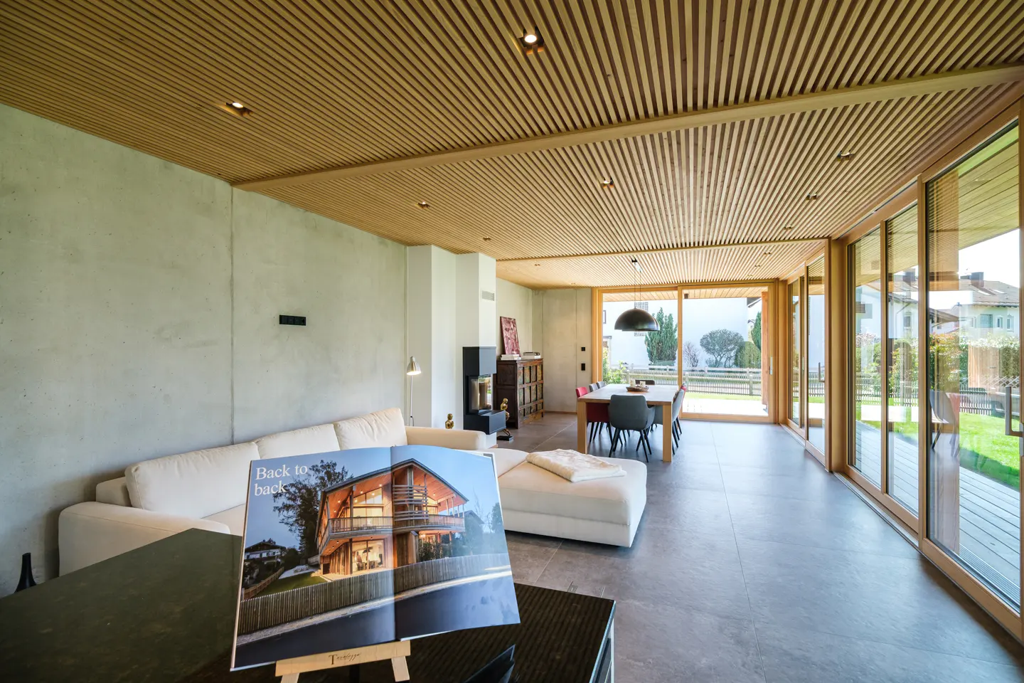 Bright living room with wood-slat ceiling, concrete walls, and floor-to-ceiling windows. A brochure displays a modern house.