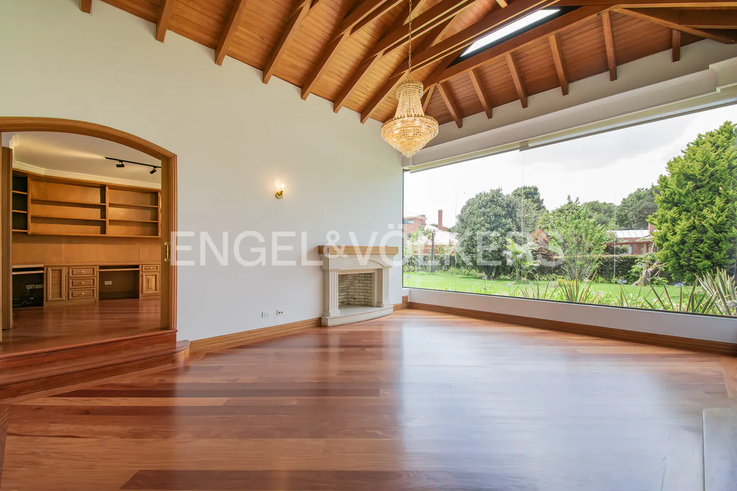 Bright living room with wood floors, a fireplace, and a large window overlooking a green lawn. A chandelier hangs from the wood ceiling.