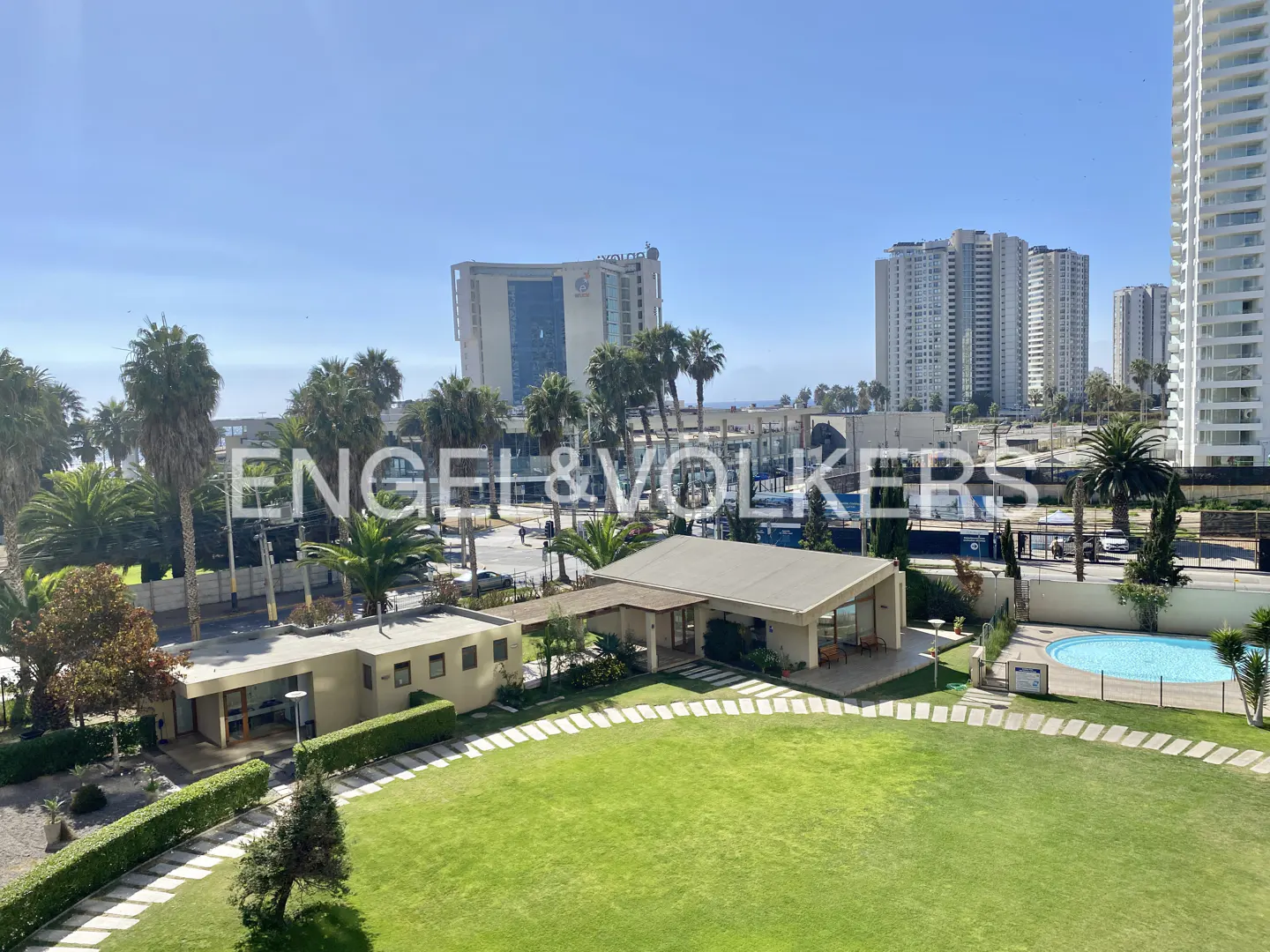 Real estate view of a green lawn, pool, and buildings under a blue sky. Palm trees are scattered throughout the landscape.