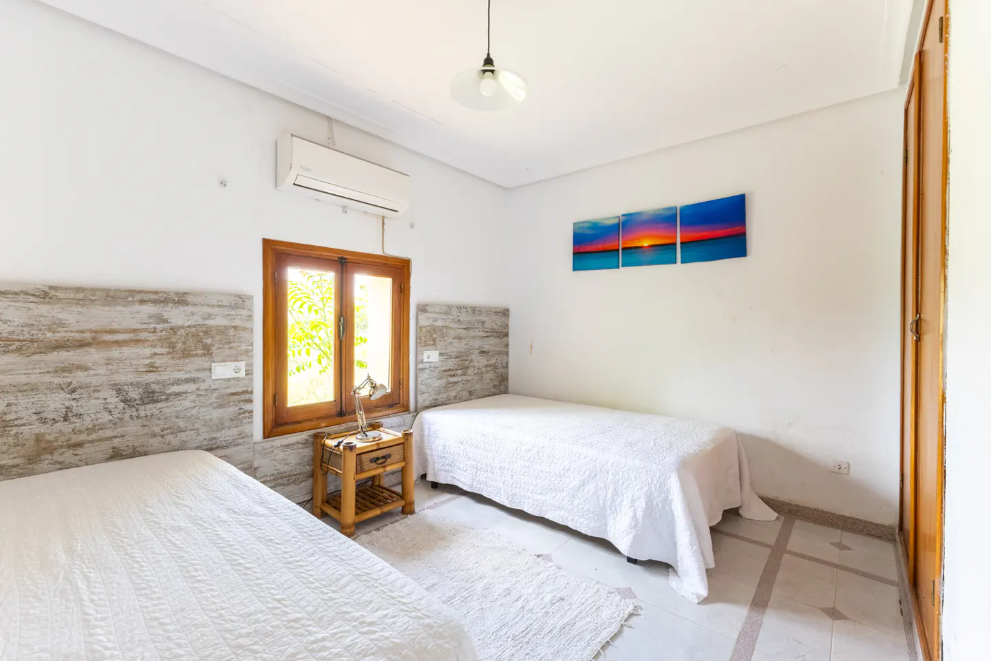 Bright bedroom with two twin beds, white walls, wood trim window, and a sunset triptych on the wall.