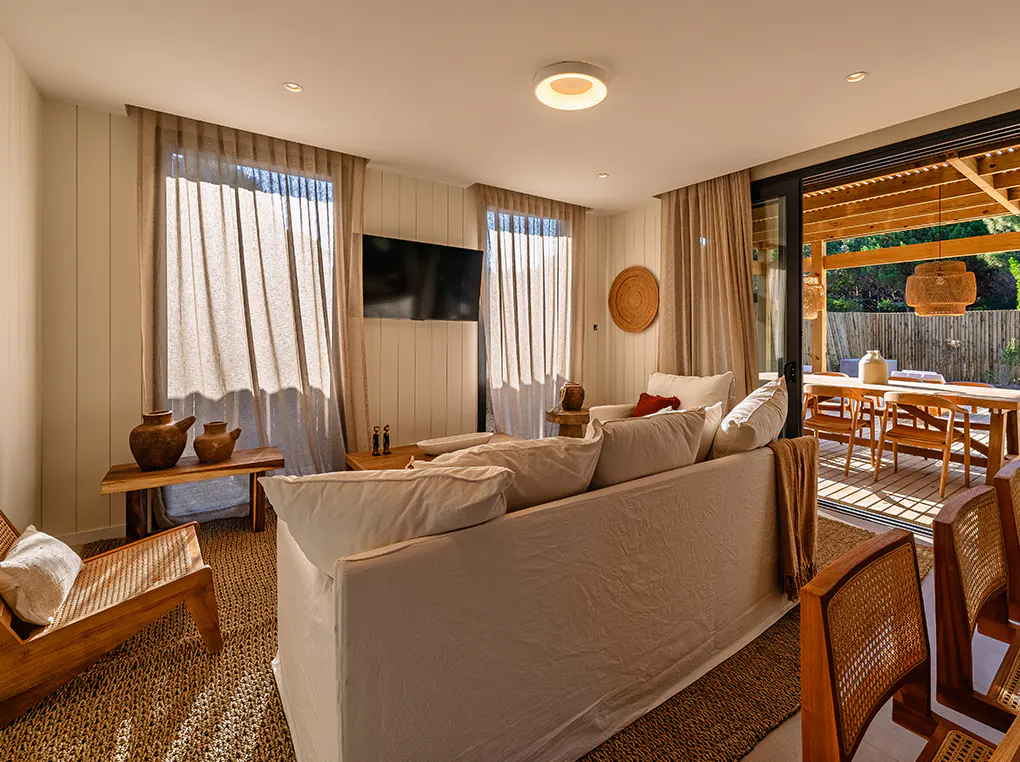 Living room with white sofa, wood furniture, and beige curtains. A dining table is visible on the patio through sliding glass doors.