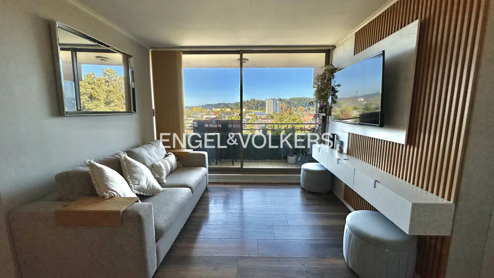 Living room with a gray sofa, wood floors, and a balcony with a city view. A TV is mounted on a wood-paneled wall.