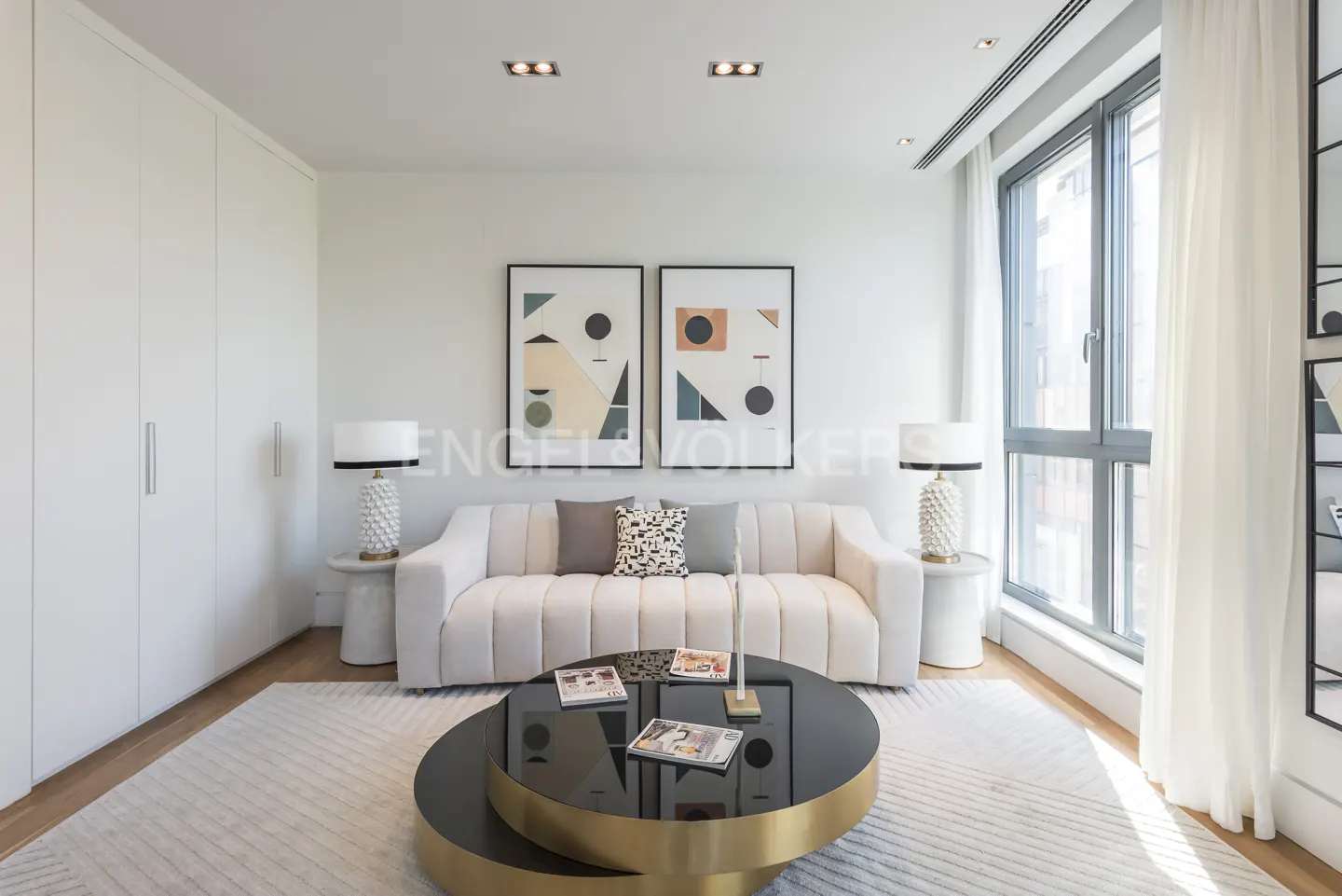 Bright living room with a white sofa, round black and gold coffee table, and abstract art on the wall. Large window with sheer curtains.