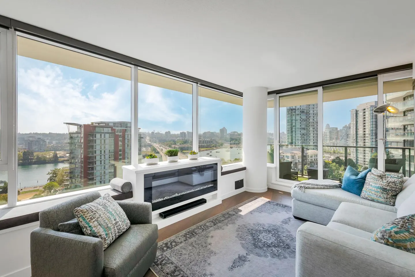 Bright living room with city view. Gray armchair, white sofa with blue pillows, and a modern fireplace. Large windows overlook city buildings.
