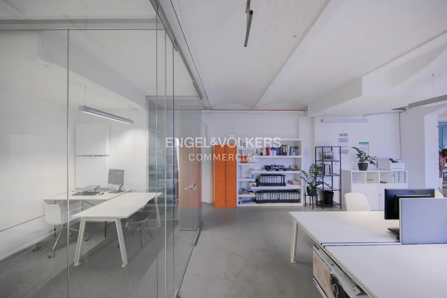 Bright office space with white walls, desks, and shelving. An orange locker and glass-walled office are visible. Engel & Völkers logo is displayed.