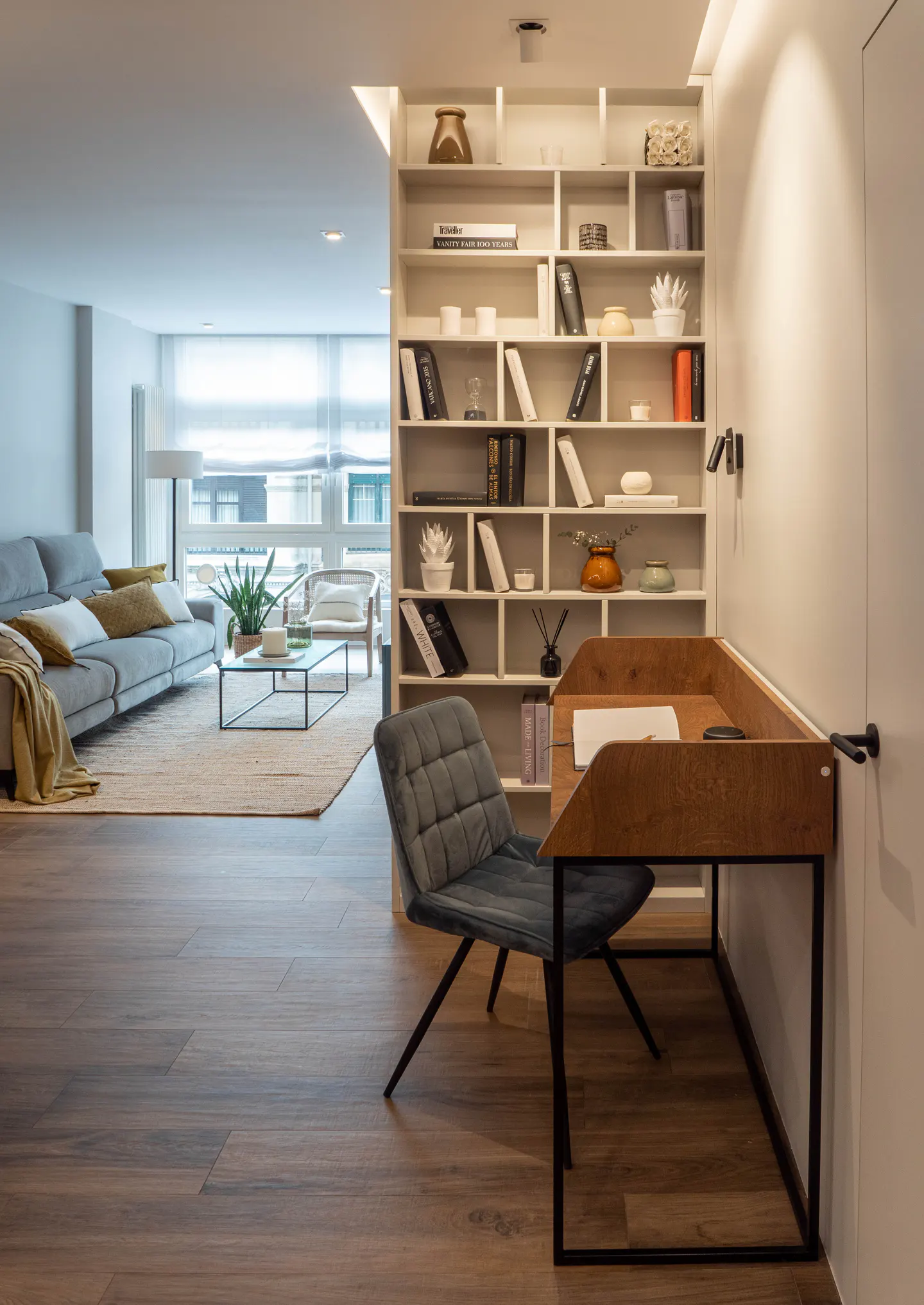A modern living room with a gray sofa, wood floors, and a desk with a gray chair. A white bookshelf is in the background.