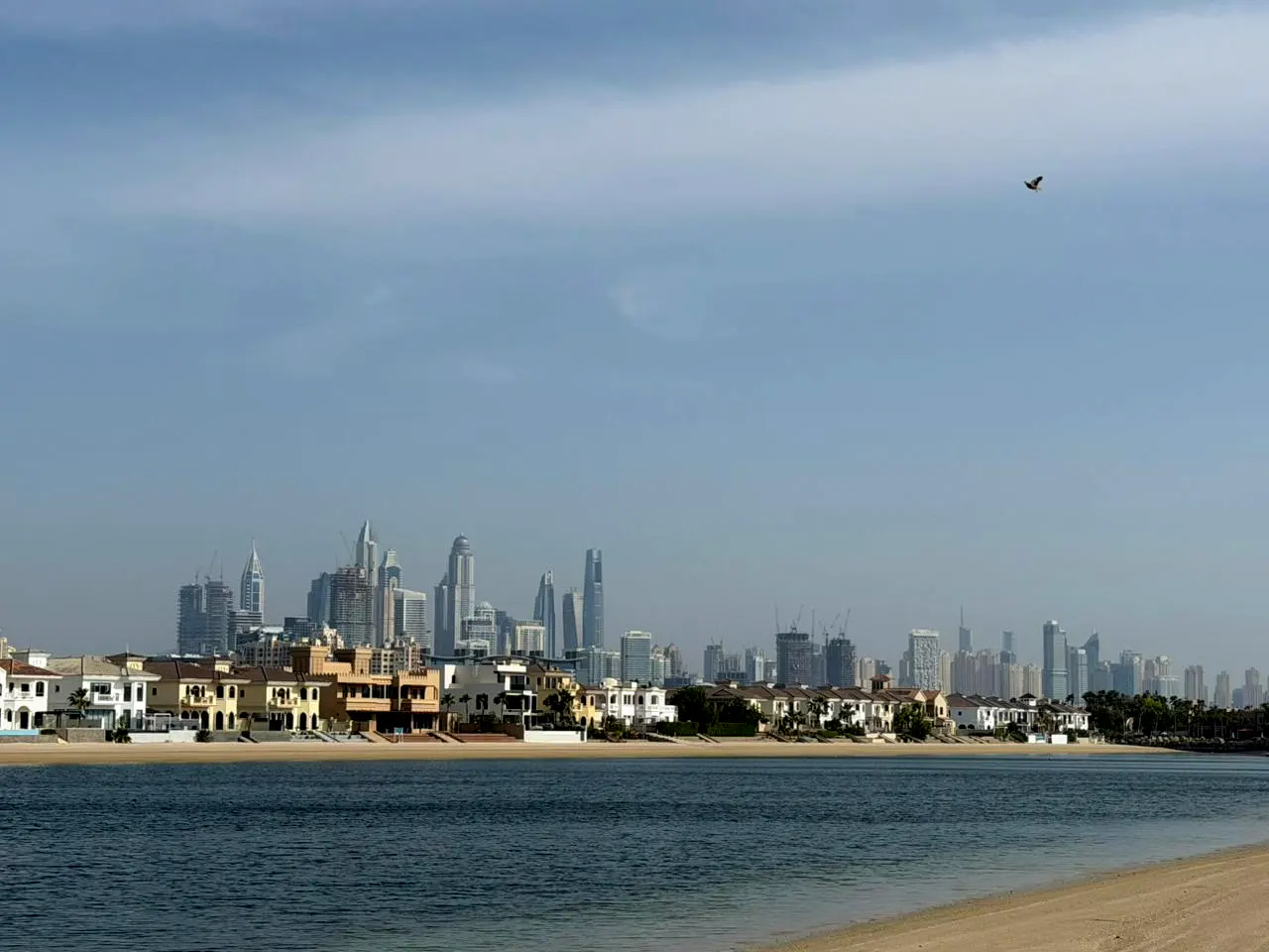 Dubai skyline view from the beach, featuring modern buildings, villas, and a bird flying in the sky.