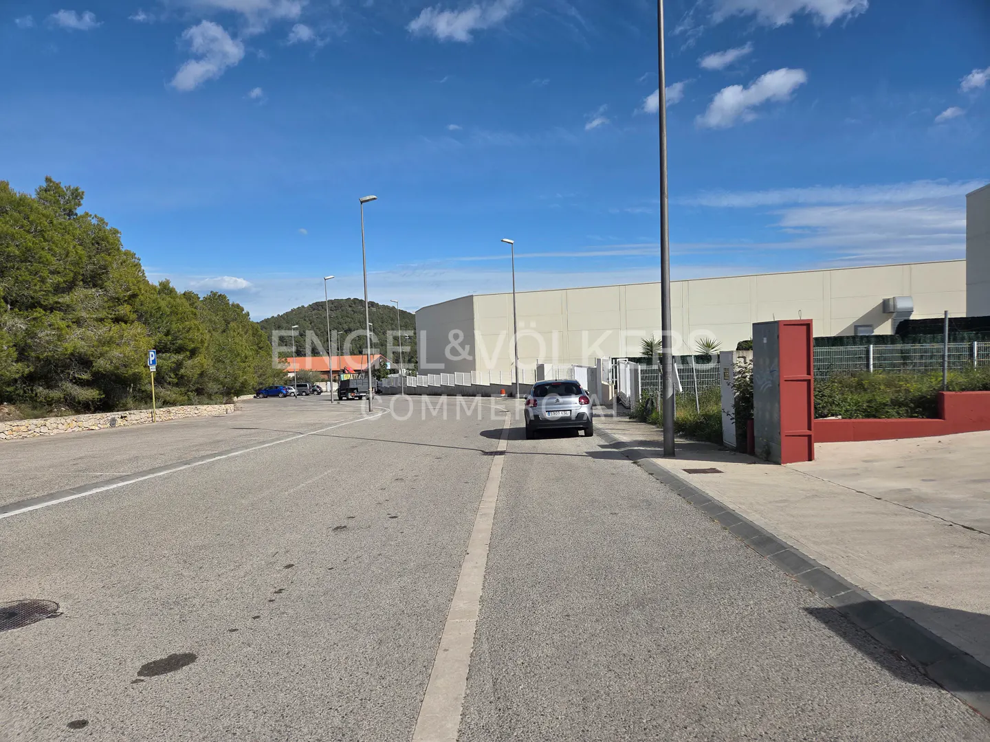 Exterior view of a gray car parked on a paved road near a large, white commercial building under a blue sky.