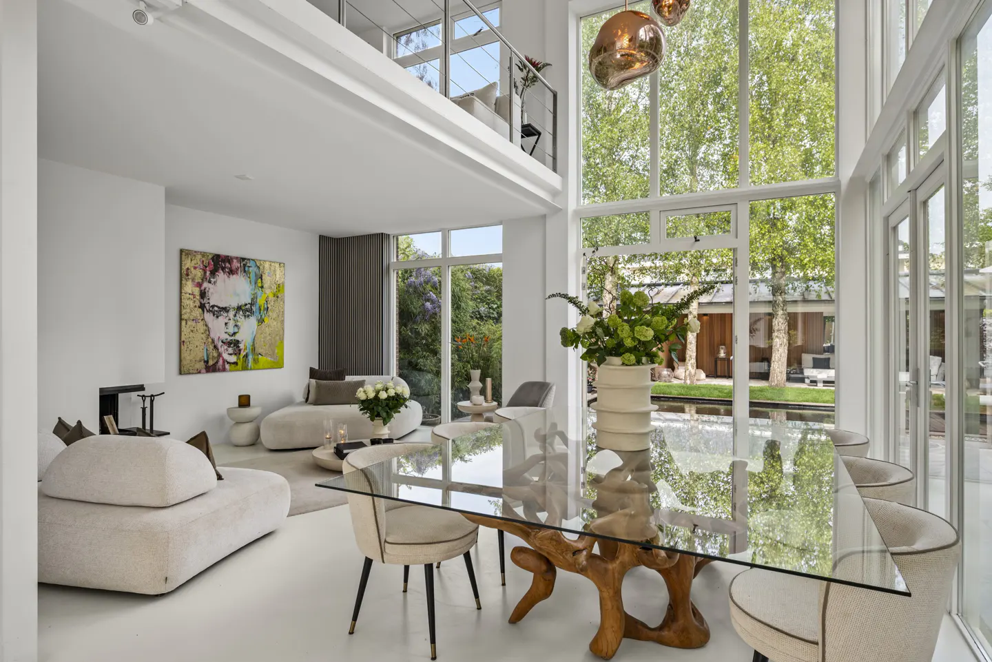 Bright, modern living room with a glass-topped table on a wood base, white sofas, and a colorful portrait. Large windows overlook a lush garden.