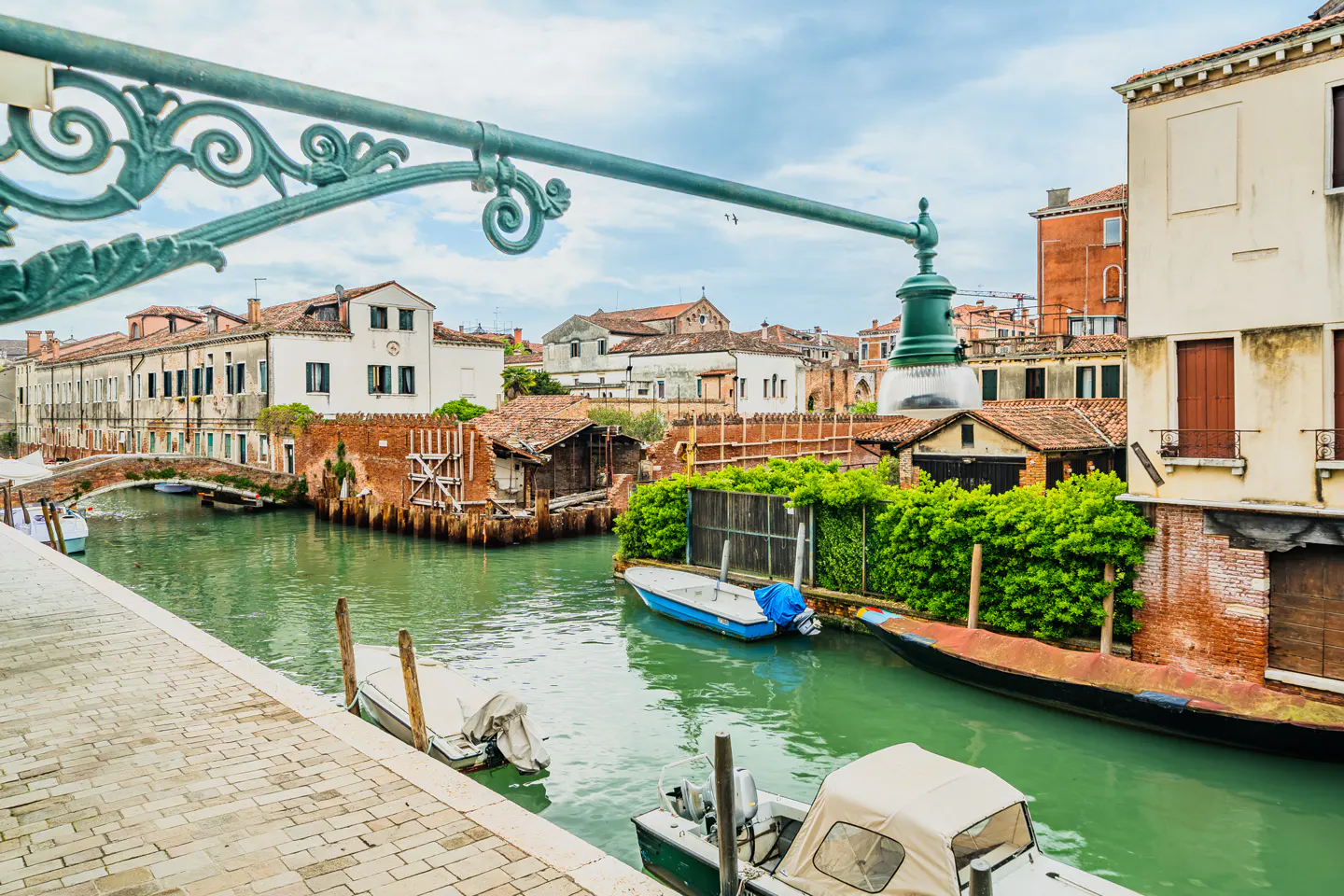 Venice canal view with boats, brick buildings, and a green ornate street lamp. The water is green and the sky is cloudy.
