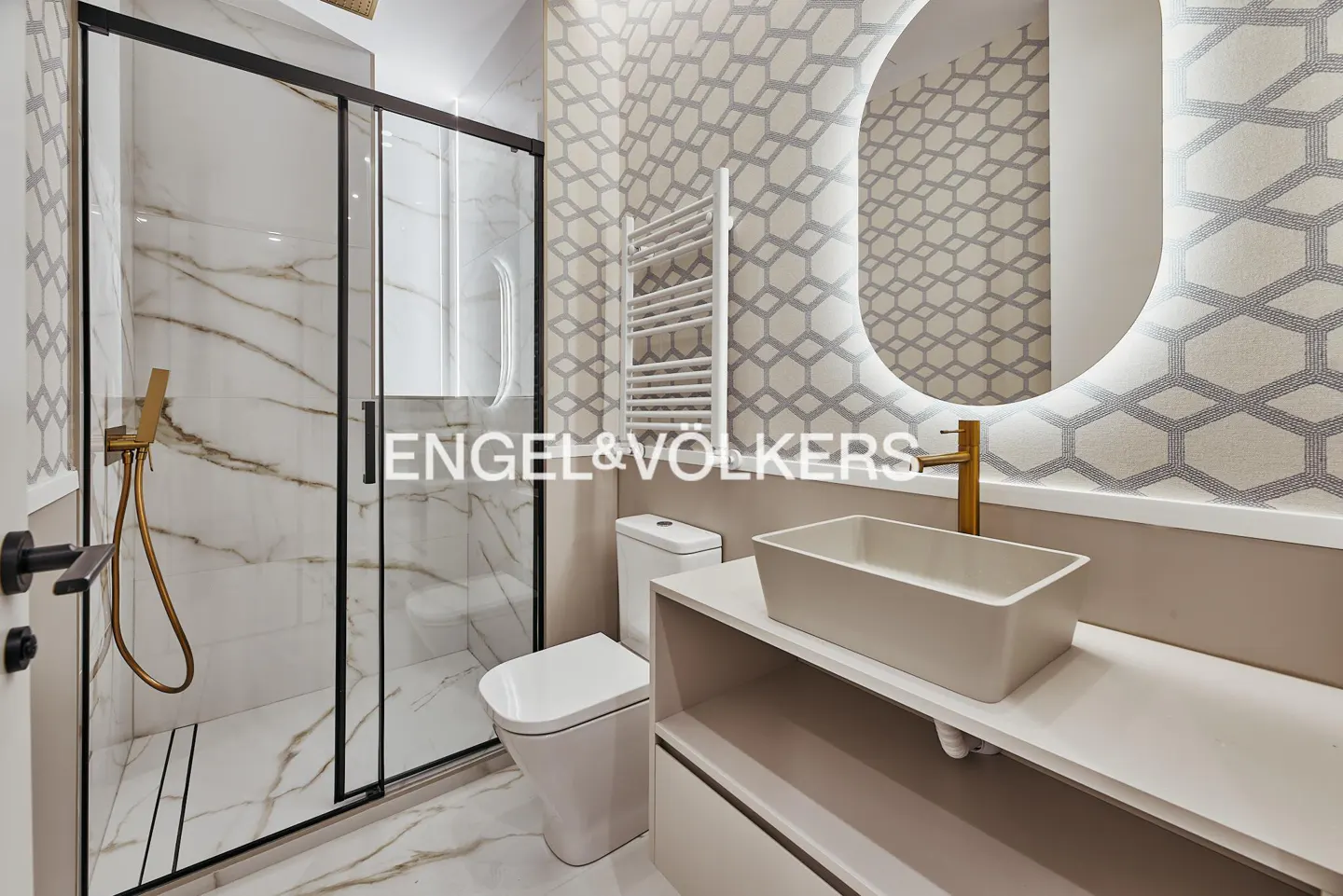 Modern bathroom with a glass shower, white toilet, sink, and a round mirror with LED lighting. The walls have a geometric pattern.