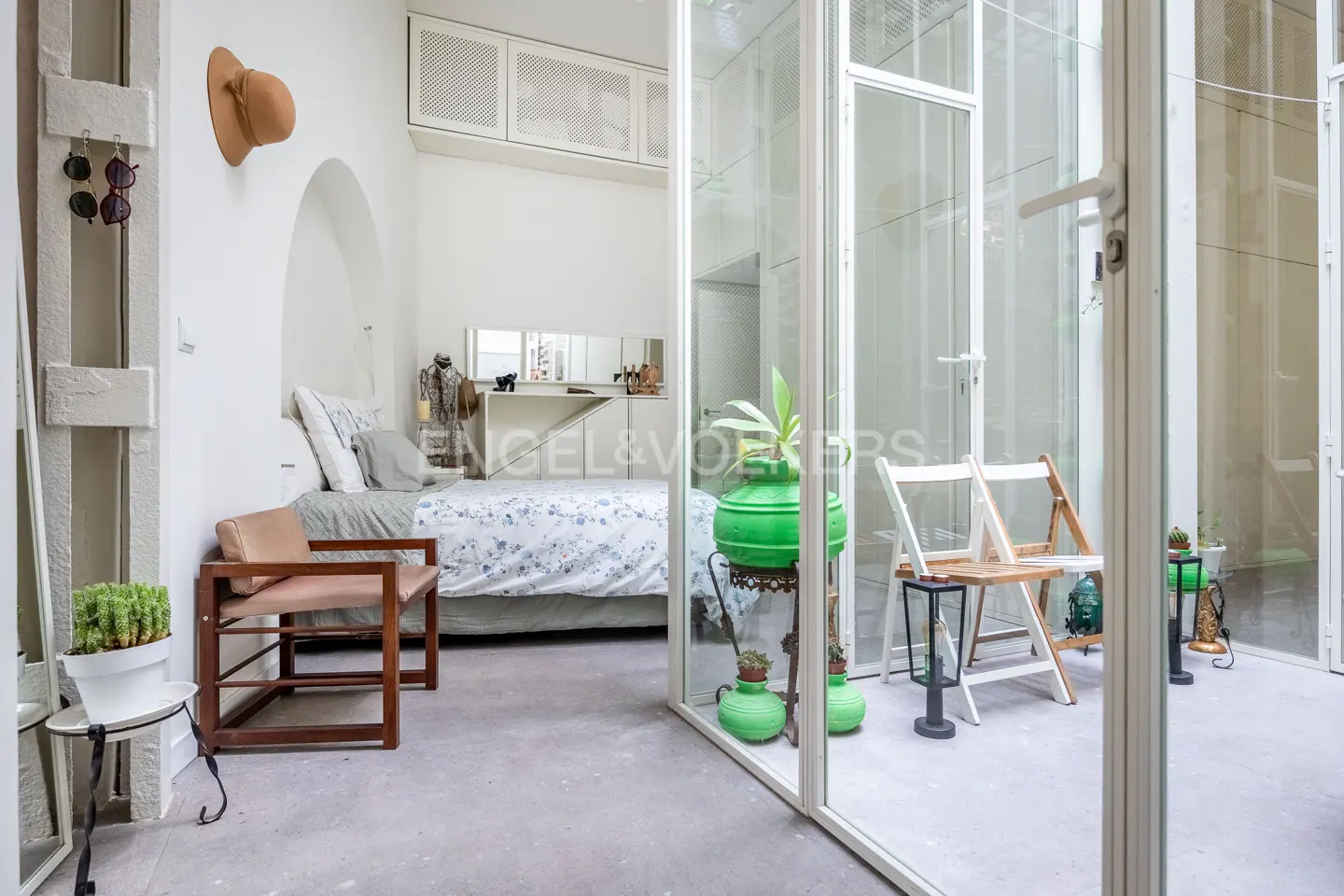 A bright bedroom with a bed, chair, and hat on the wall, seen through glass doors.