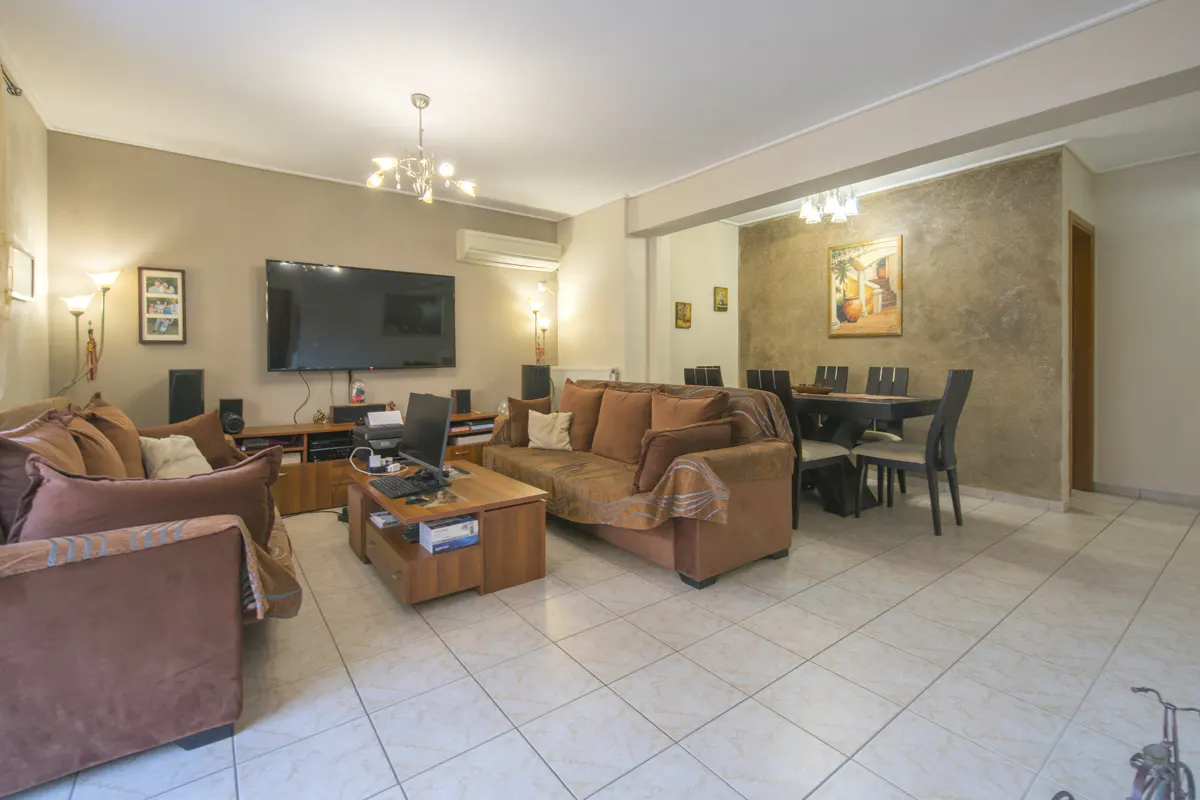 A living room with brown sofas, a TV, a dining table, and tiled floors.