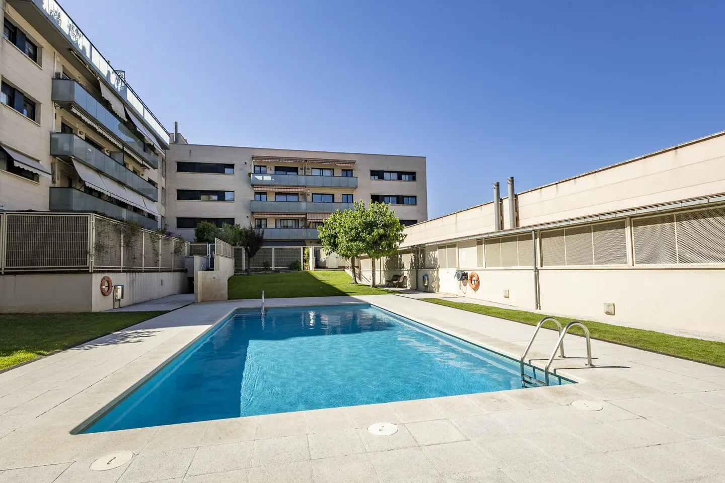 Rectangular swimming pool with blue water, surrounded by a stone patio, green grass, and modern apartment buildings.