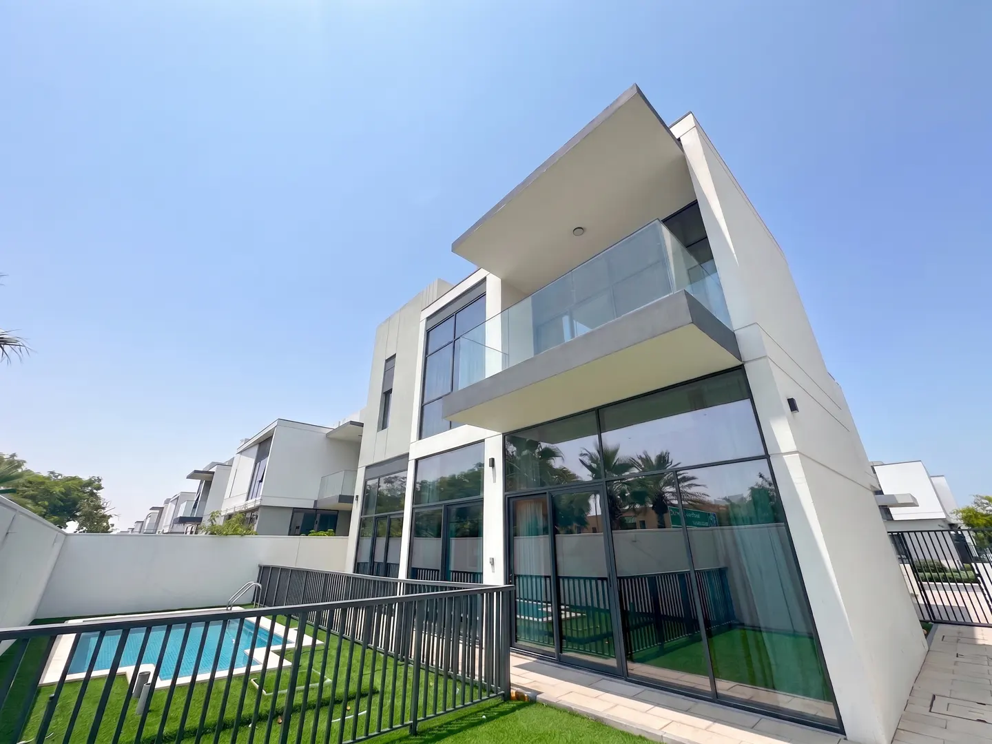 Modern two-story white house with a balcony and large windows, overlooking a pool and green lawn.
