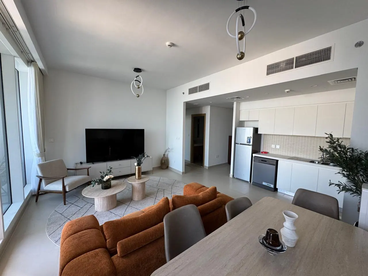 Bright, modern apartment interior with an orange sofa, dining table, TV, and white kitchen. Large windows provide natural light.