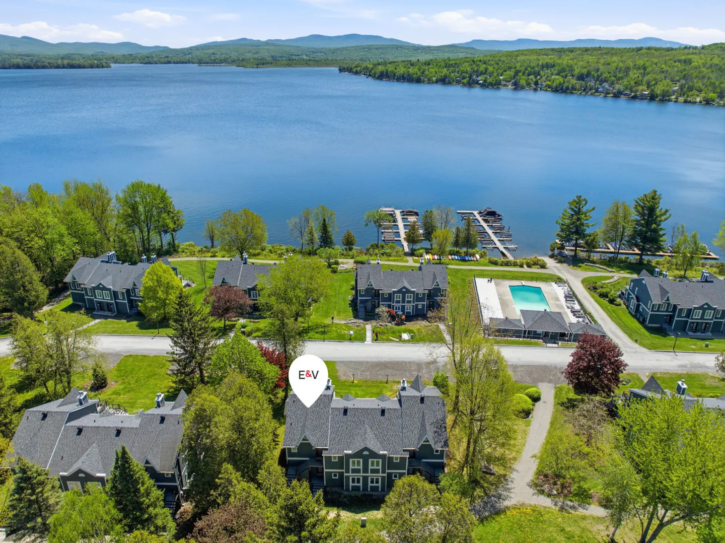 Aerial view of lakeside condos with a pool and boat docks on a sunny day. Green trees surround the buildings.