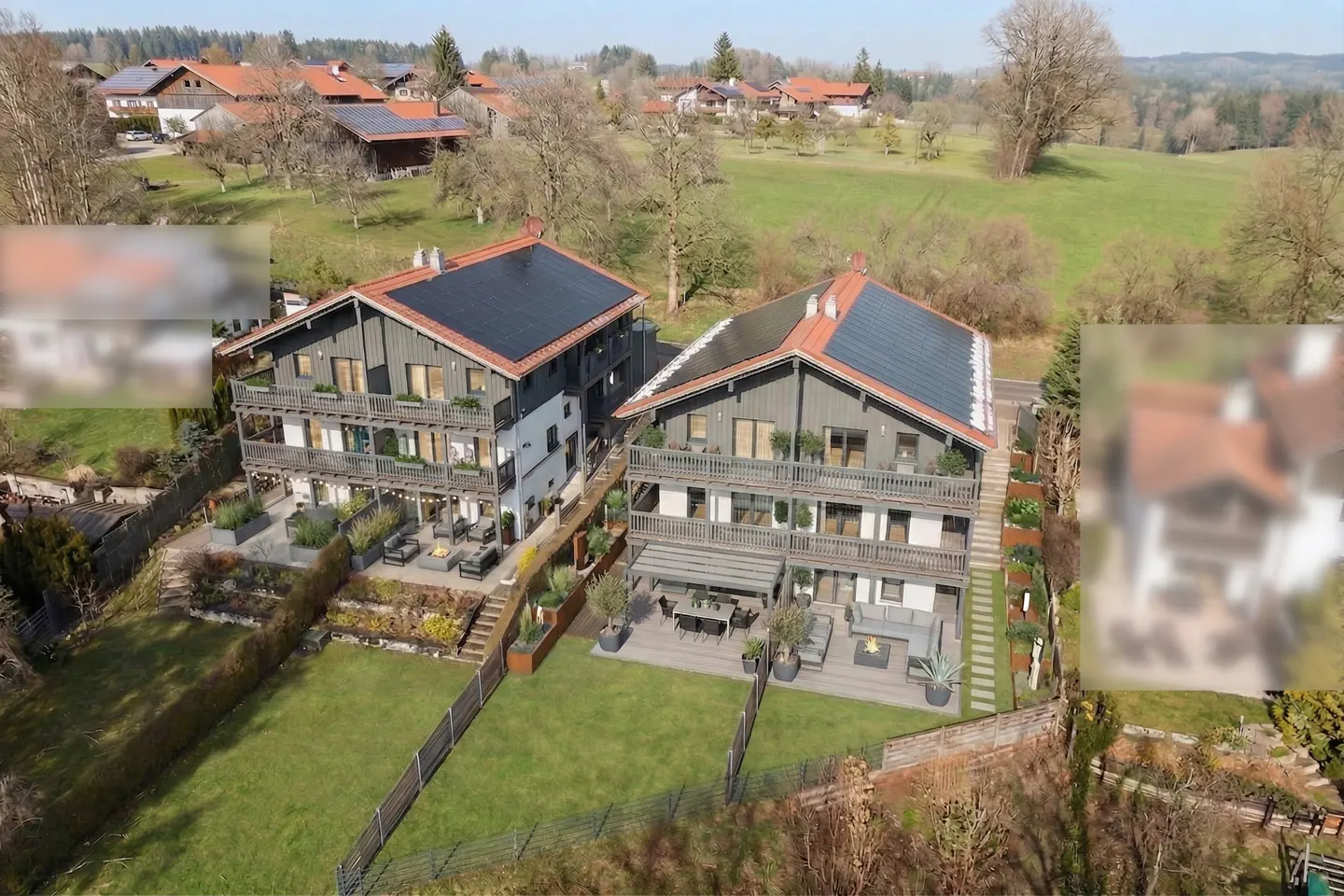 Aerial view of two modern, three-story homes with dark gray siding, balconies, and solar panels, set in a green, rural landscape.
