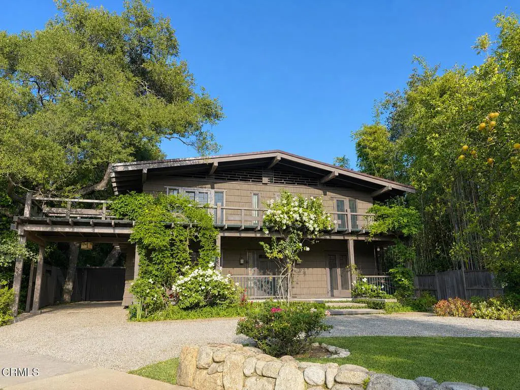 Two-story brown house with a balcony, surrounded by lush greenery and a gravel driveway. Blue sky in the background.