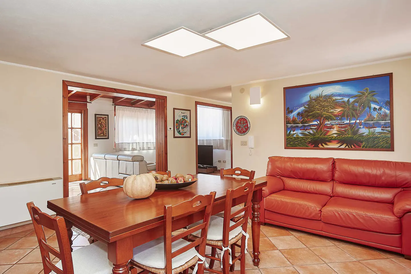 A dining room with a wooden table, chairs, and a red leather sofa. A painting of palm trees hangs on the wall.