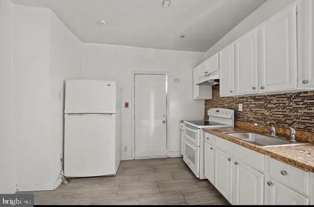 A bright kitchen with white cabinets, appliances, and walls. A brown tiled backsplash and floor add contrast.