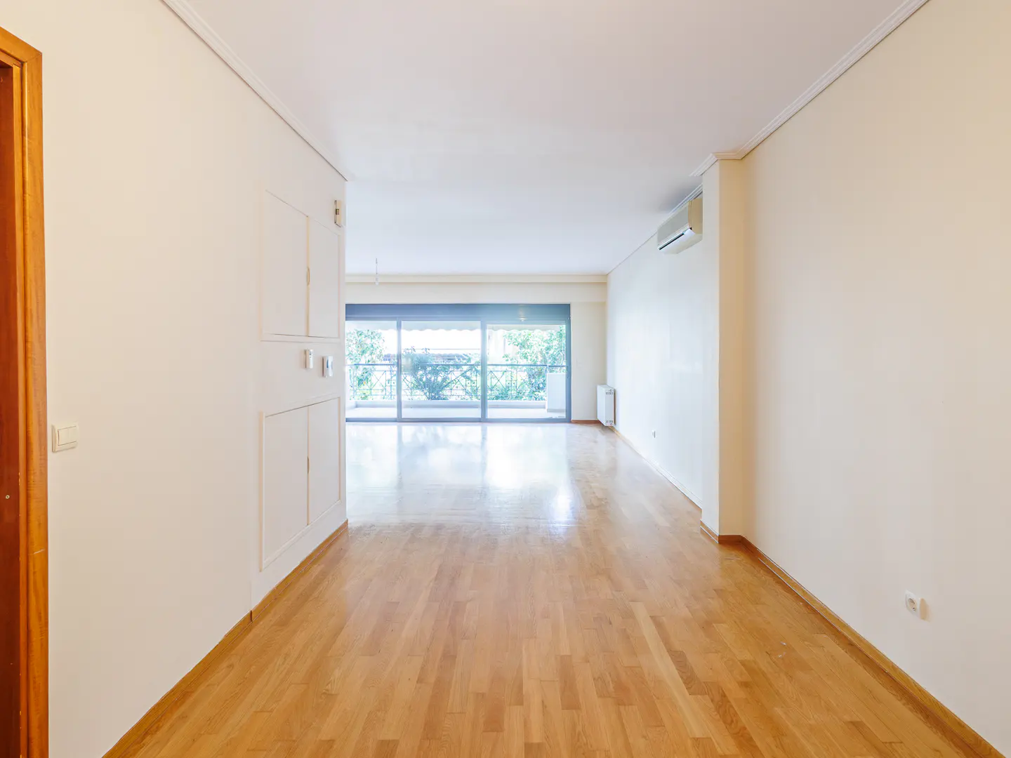 Empty room with light wood floors, cream walls, and a large sliding glass door to a balcony with green trees.