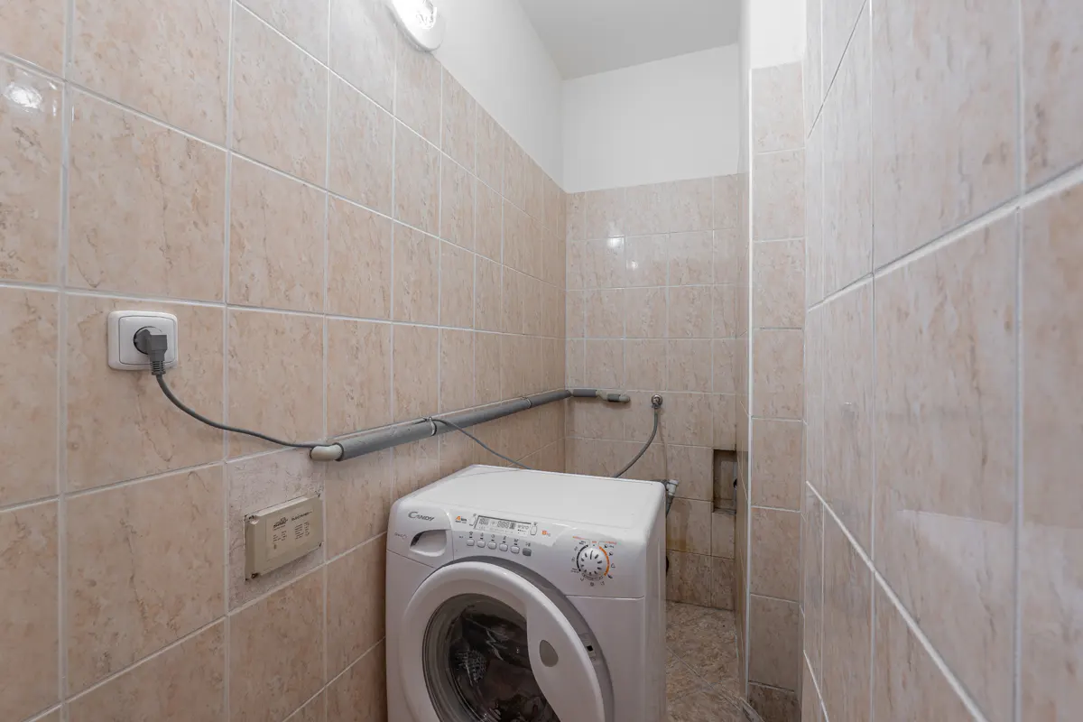 A laundry room with beige tiled walls features a white Candy washing machine, grey pipes, and an electrical outlet.