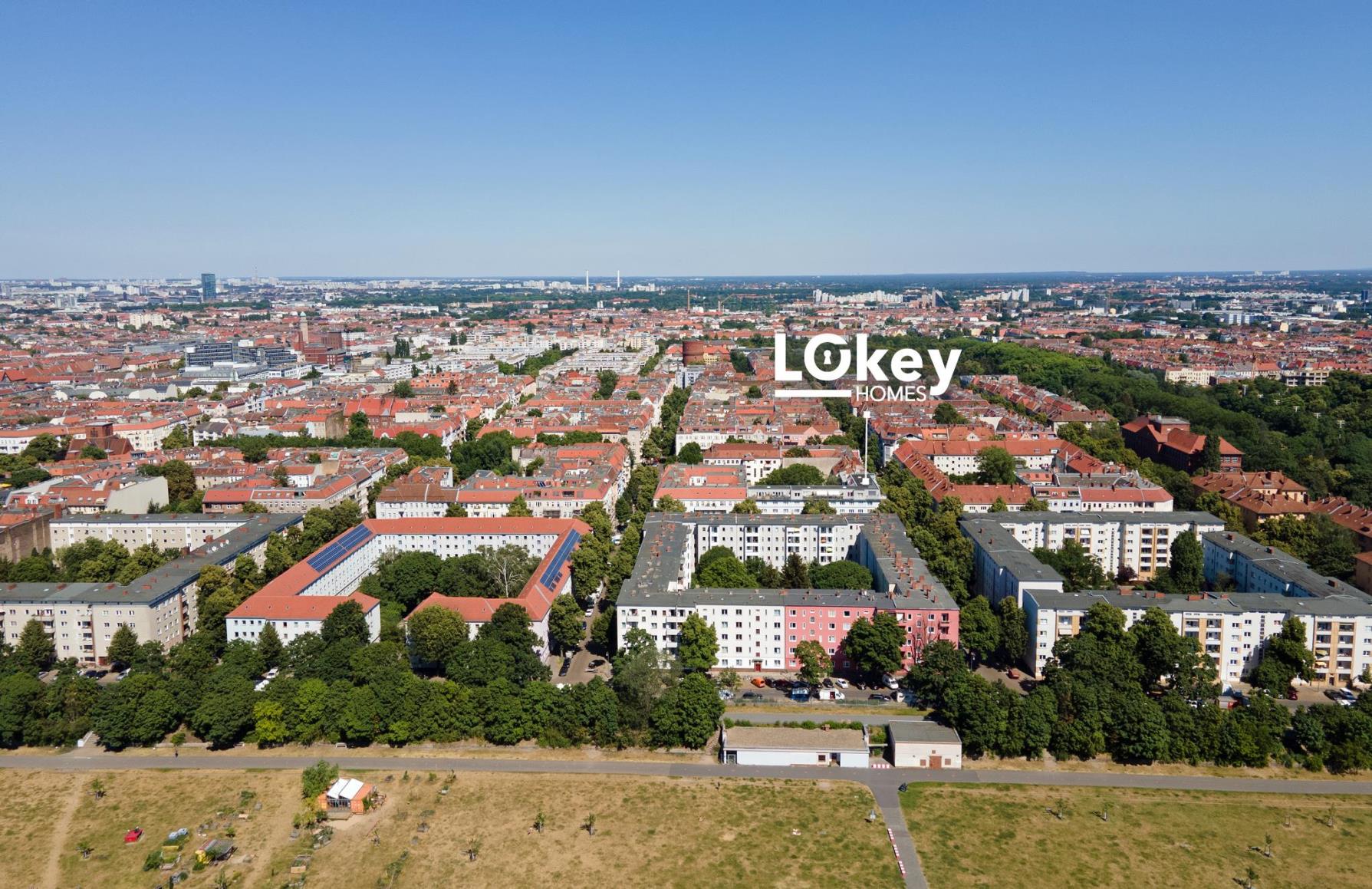 Aerial view of a city neighborhood with apartment blocks, green trees, and a clear blue sky, featuring the Lokey Homes logo. [AI-generated text]