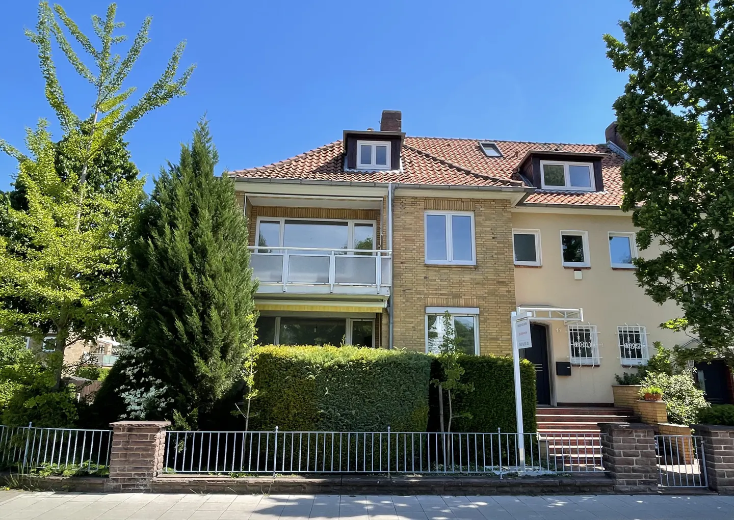 Two-story brick house with a red tile roof and white trim, surrounded by green trees and a metal fence.