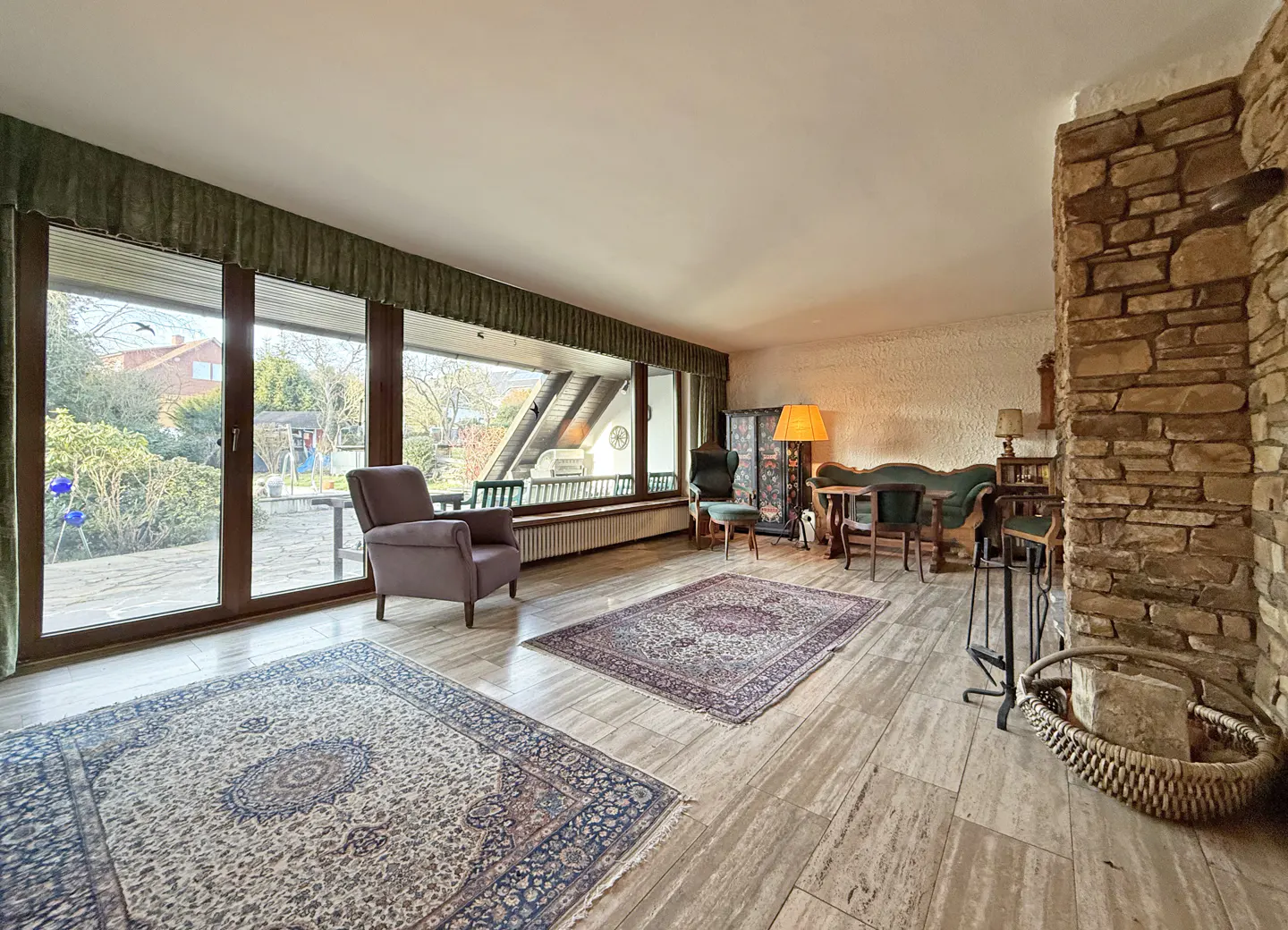 A living room with large windows, a stone wall, and two patterned rugs on a light-colored floor. A purple armchair sits near the windows.