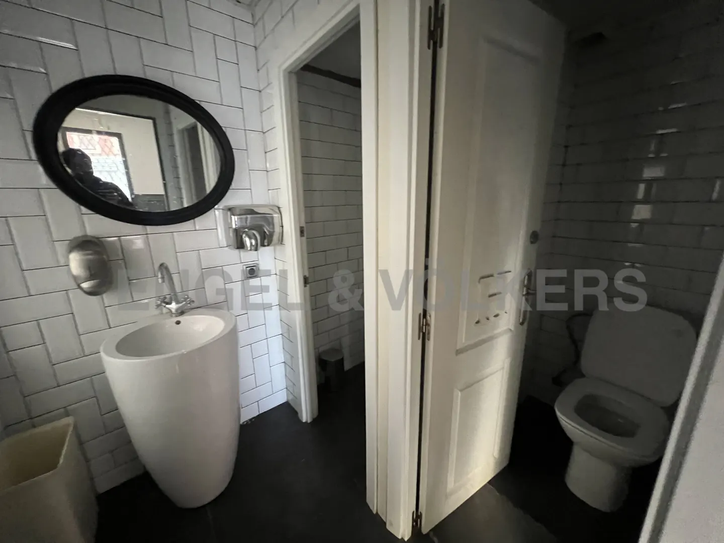 A modern bathroom with white subway tile, a pedestal sink, and a black oval mirror. A toilet is visible through an open door.