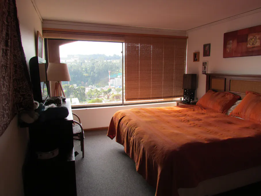 Bedroom with orange bedding, a large window with bamboo blinds, and a view of a city and trees.