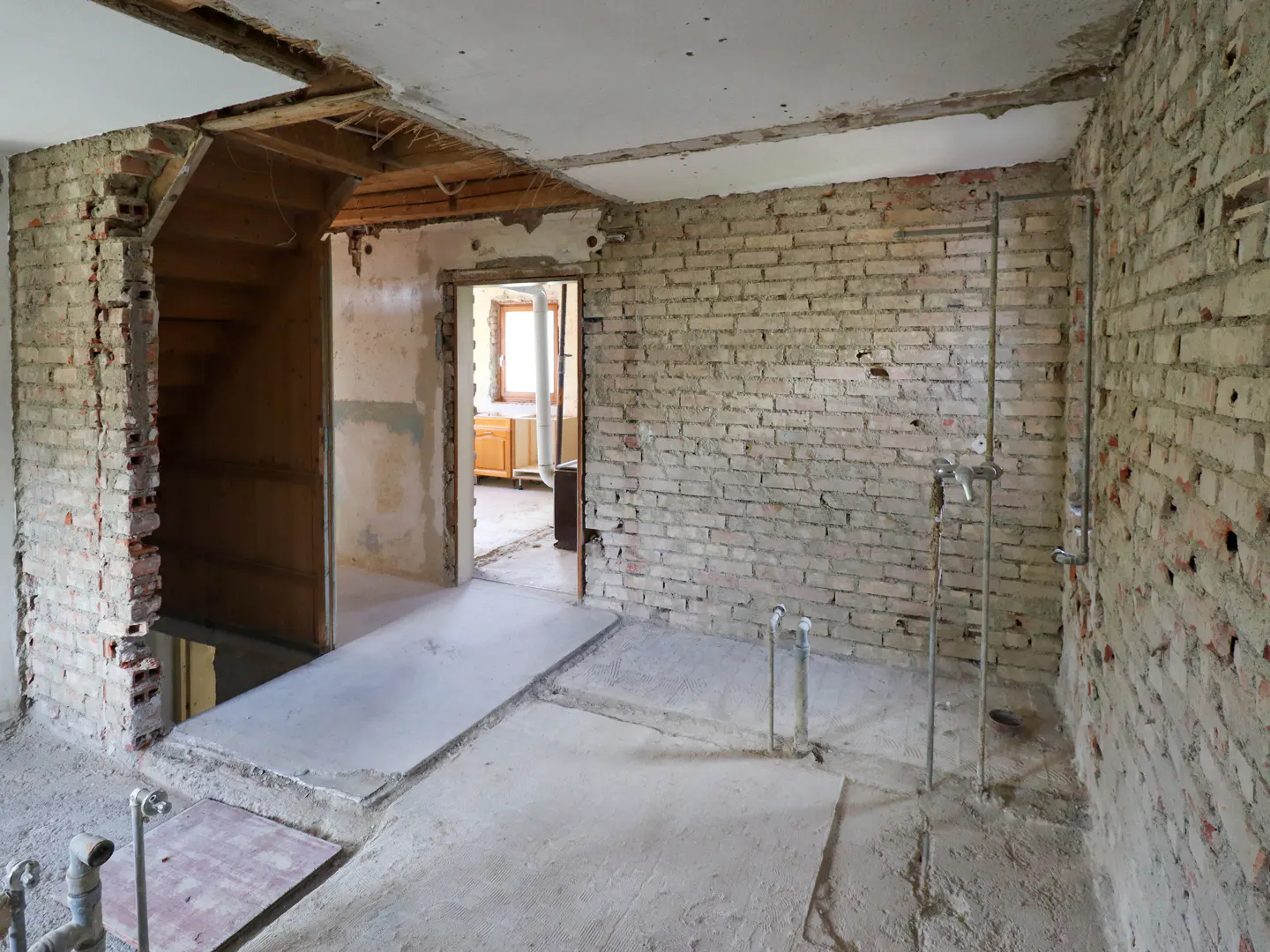 Interior view of a room under renovation with exposed brick walls, concrete floors, and visible plumbing. A staircase and doorway lead to other rooms.