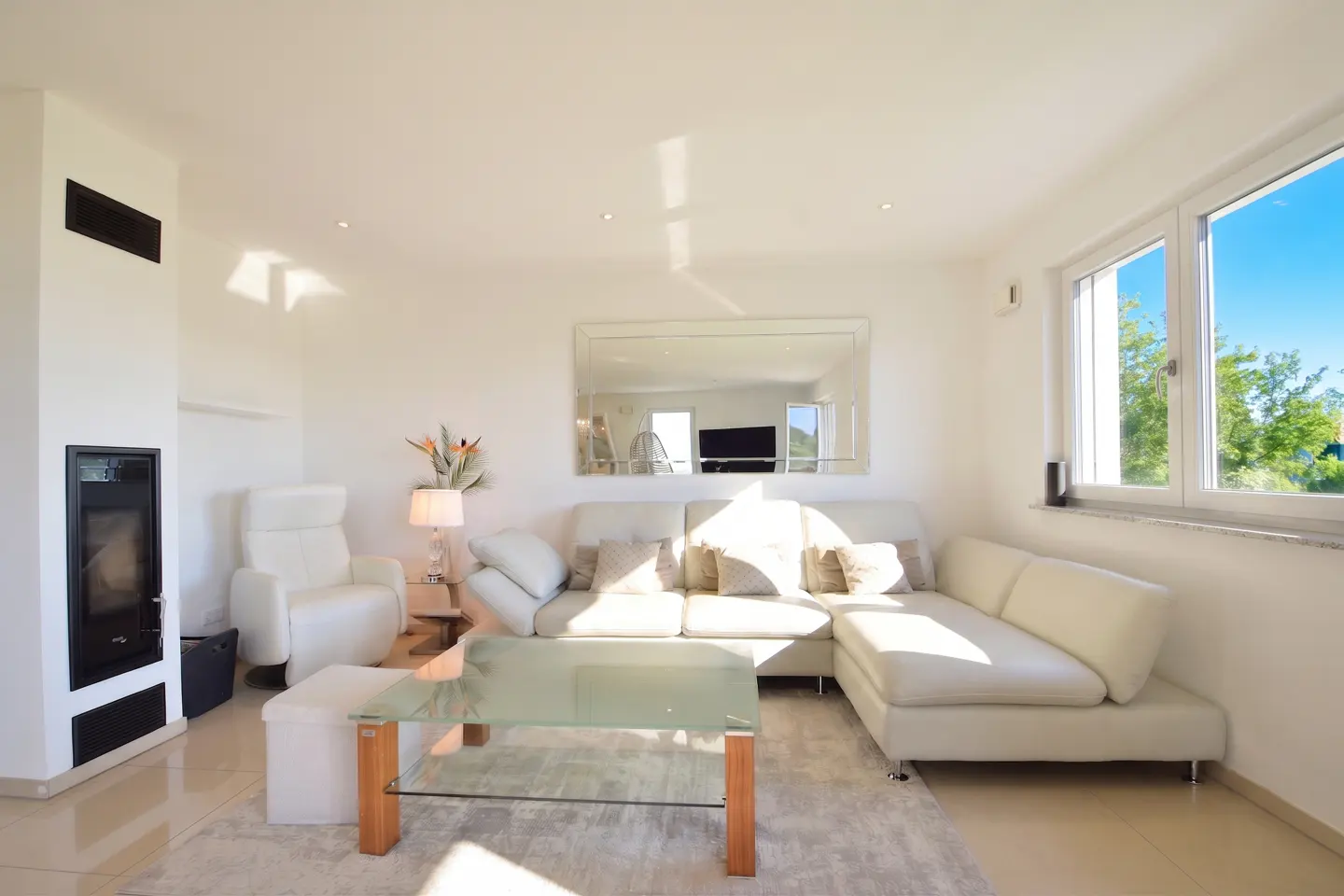 Bright living room with white walls, a fireplace, and a large window with a view of trees and blue sky. A white sectional sofa and glass coffee table sit on a light gray rug.