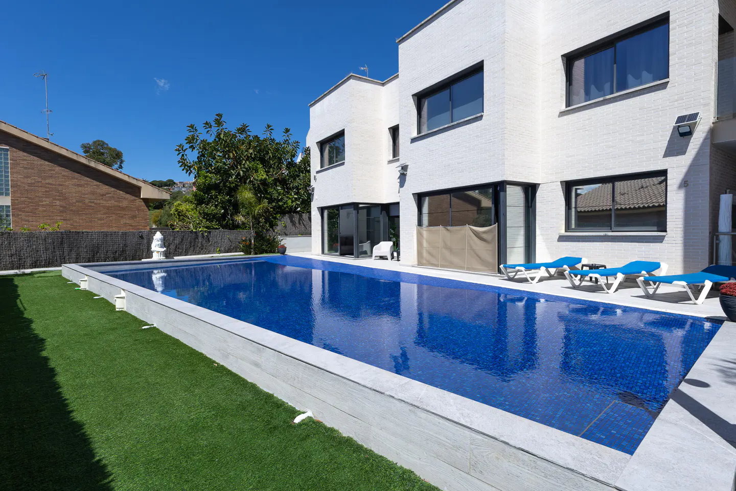 A modern white house with a blue tiled pool, green grass, and blue lounge chairs under a clear blue sky.
