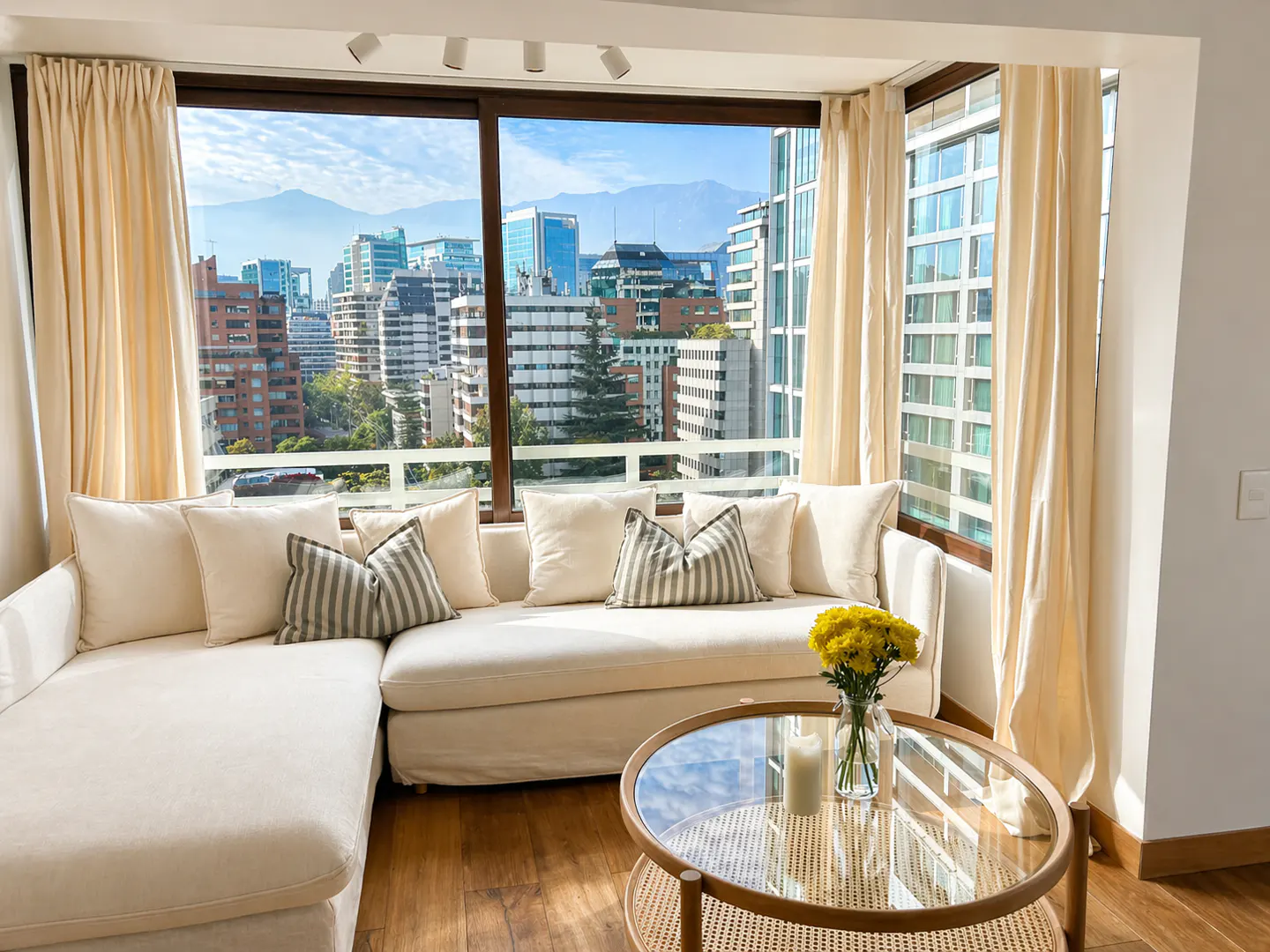 Bright living room with a white sectional sofa, striped pillows, and a glass coffee table with yellow flowers, overlooking a city skyline.