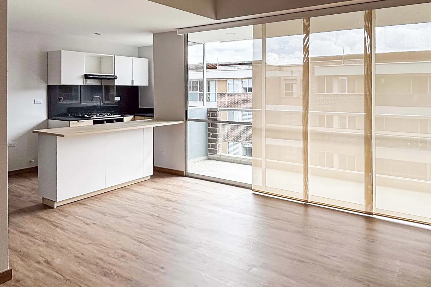 An open-concept apartment with a white kitchen island, cabinets, and a sliding glass door to a balcony. The floor is light brown wood.