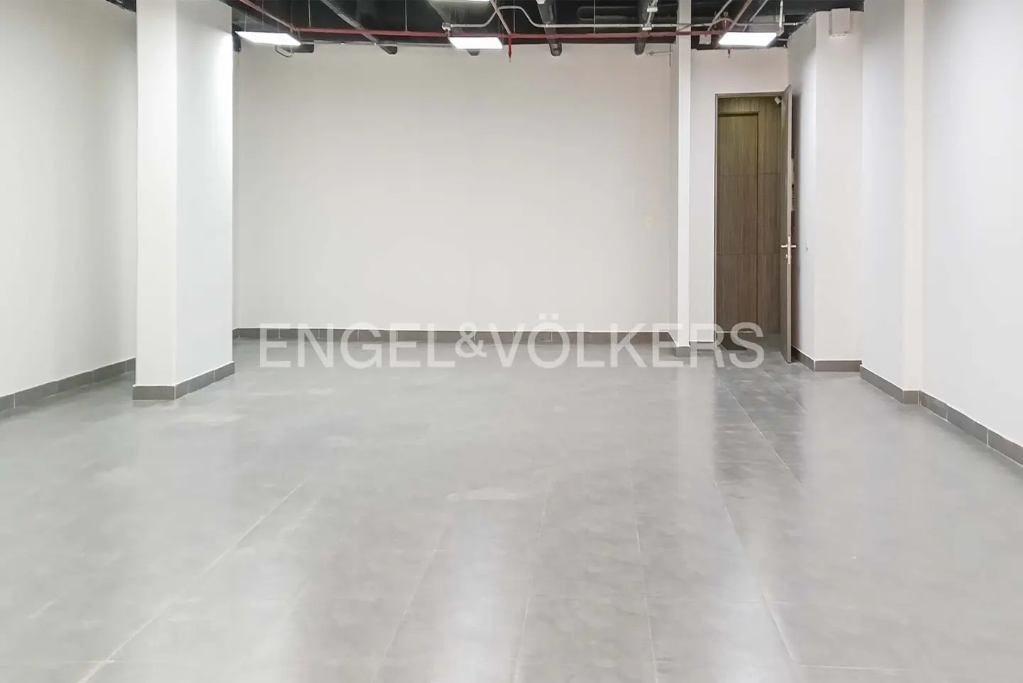 Empty commercial space with gray tile floor, white walls, and a brown door. Overhead pipes and lights are visible.
