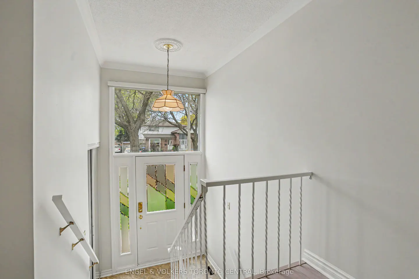 Interior view of a white entryway with a stained glass door, white railing, and a hanging light fixture. A tree is visible through the door.