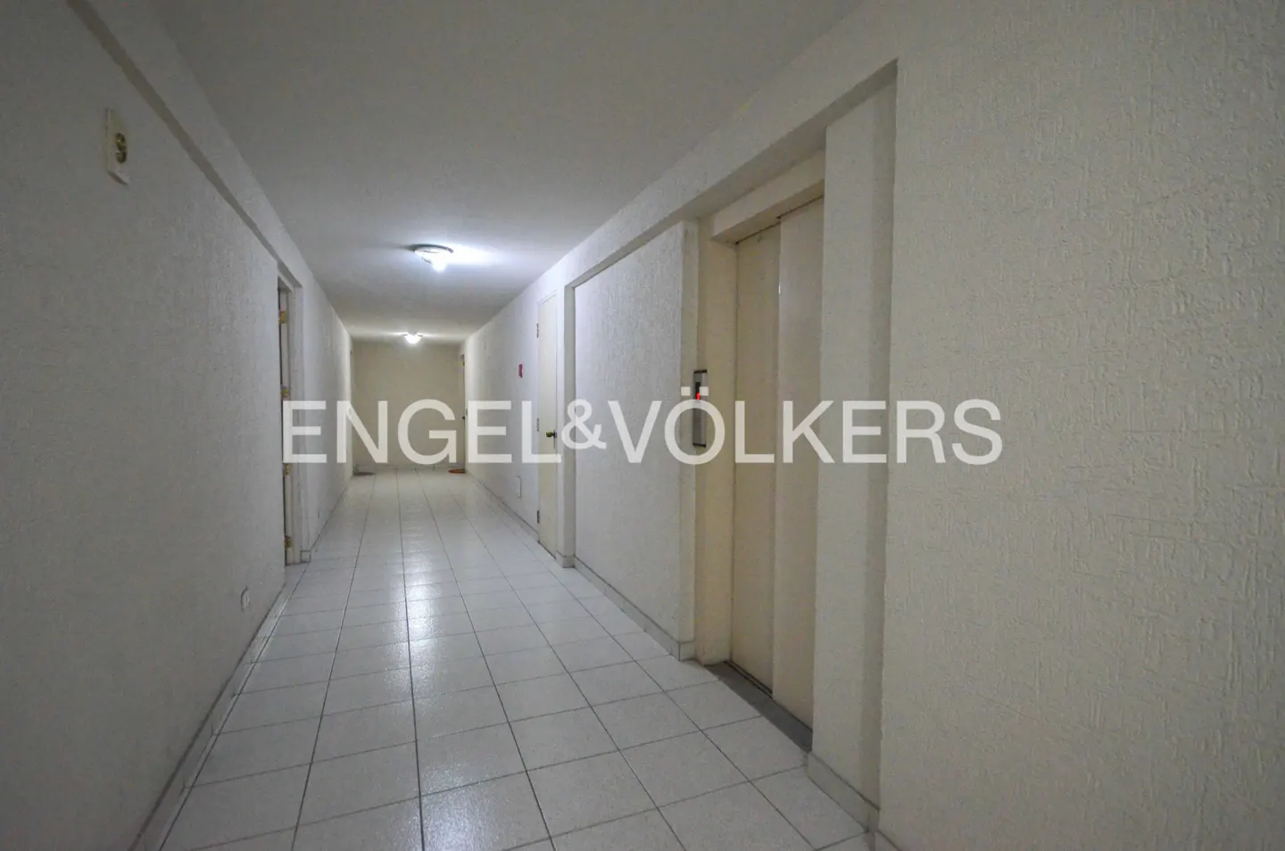 Long, empty hallway with white tile floors and walls, doors on both sides, and recessed lighting.