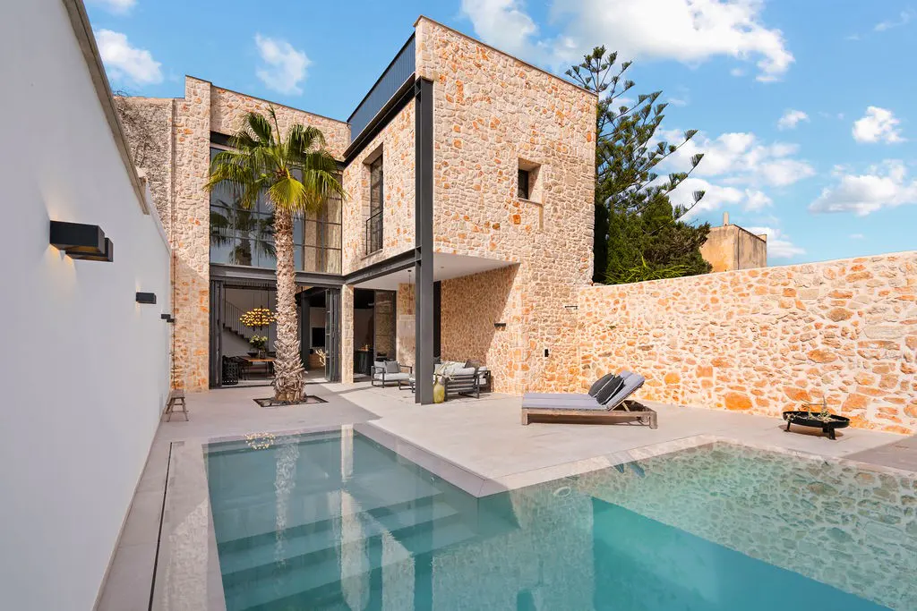 Exterior view of a stone house with a pool, lounge chairs, and a palm tree.