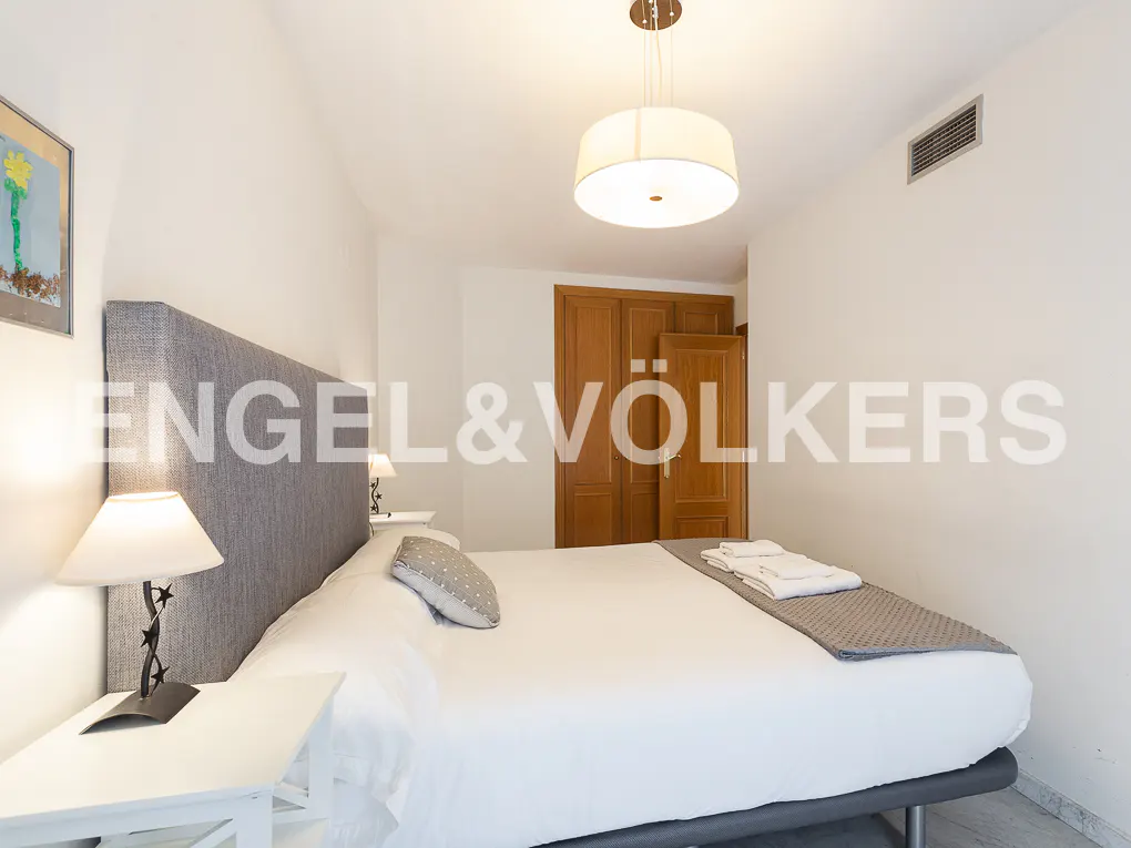 Bedroom with a gray headboard, white linens, and a drum pendant light. A wooden wardrobe is in the background.