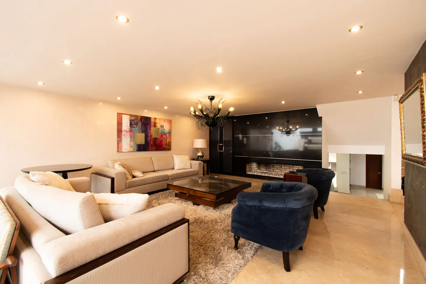 Bright living room with beige sofas, blue chairs, and a black fireplace. A colorful painting hangs on the wall.